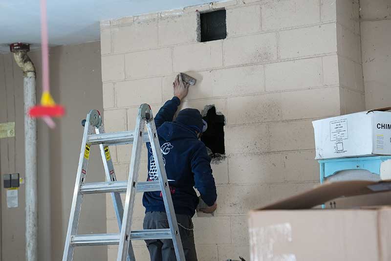 Tech inspecting hole in masonry wall with ladder behind him.