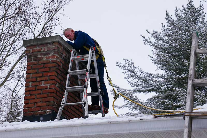 Tech looking down chimney using ladder and safety equipment on a snow covered roof.